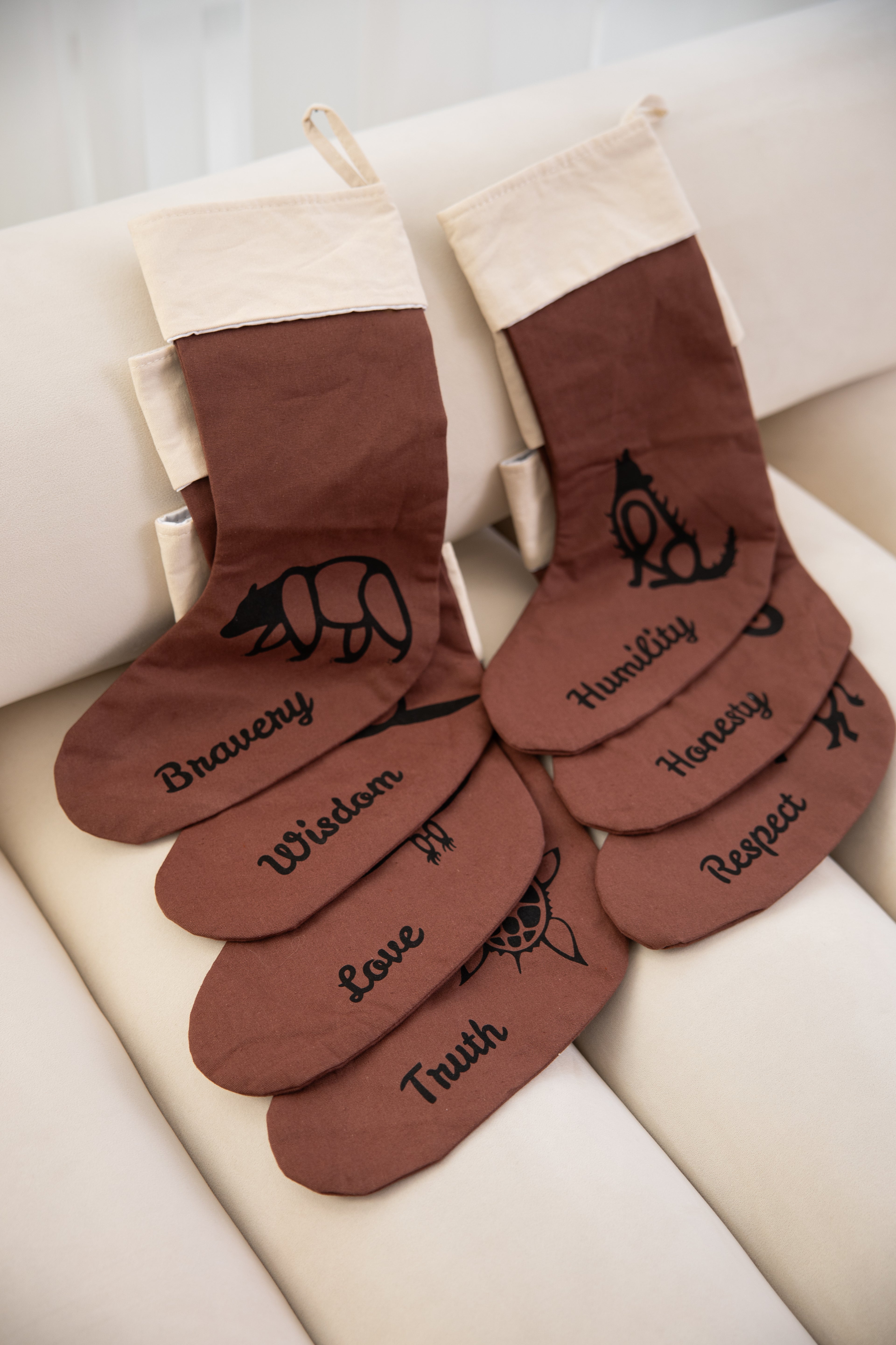 Brown Christmas stockings with embroidered text on a white surface. The Embroidered text displays a woodland cultural entity next to one of seven Indigenous grandfather teachings.