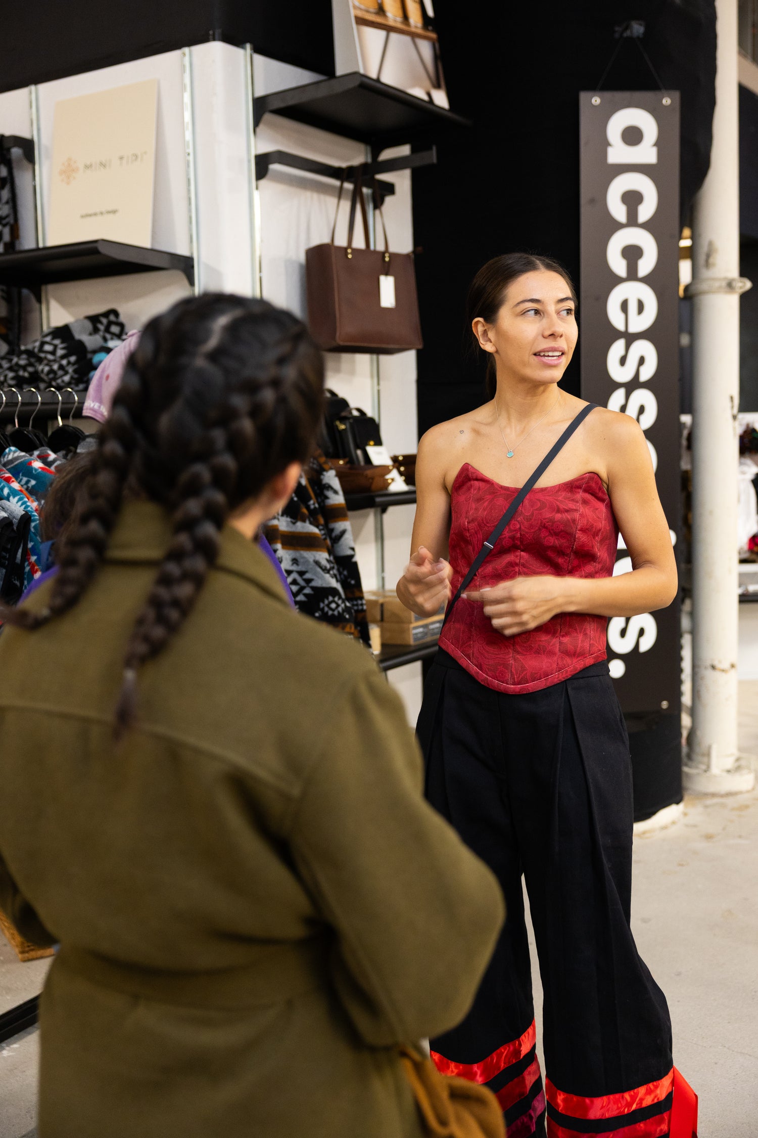 Founder and CEO Chelsee Pettit in a red strapless top and black pants trimmed with red ribbon standing in a store with a customer during a marketing event for Aaniin Apparel in the pop-up store at the Eaton Centre Mall. Photo by Benjamin's Agency Inc.
