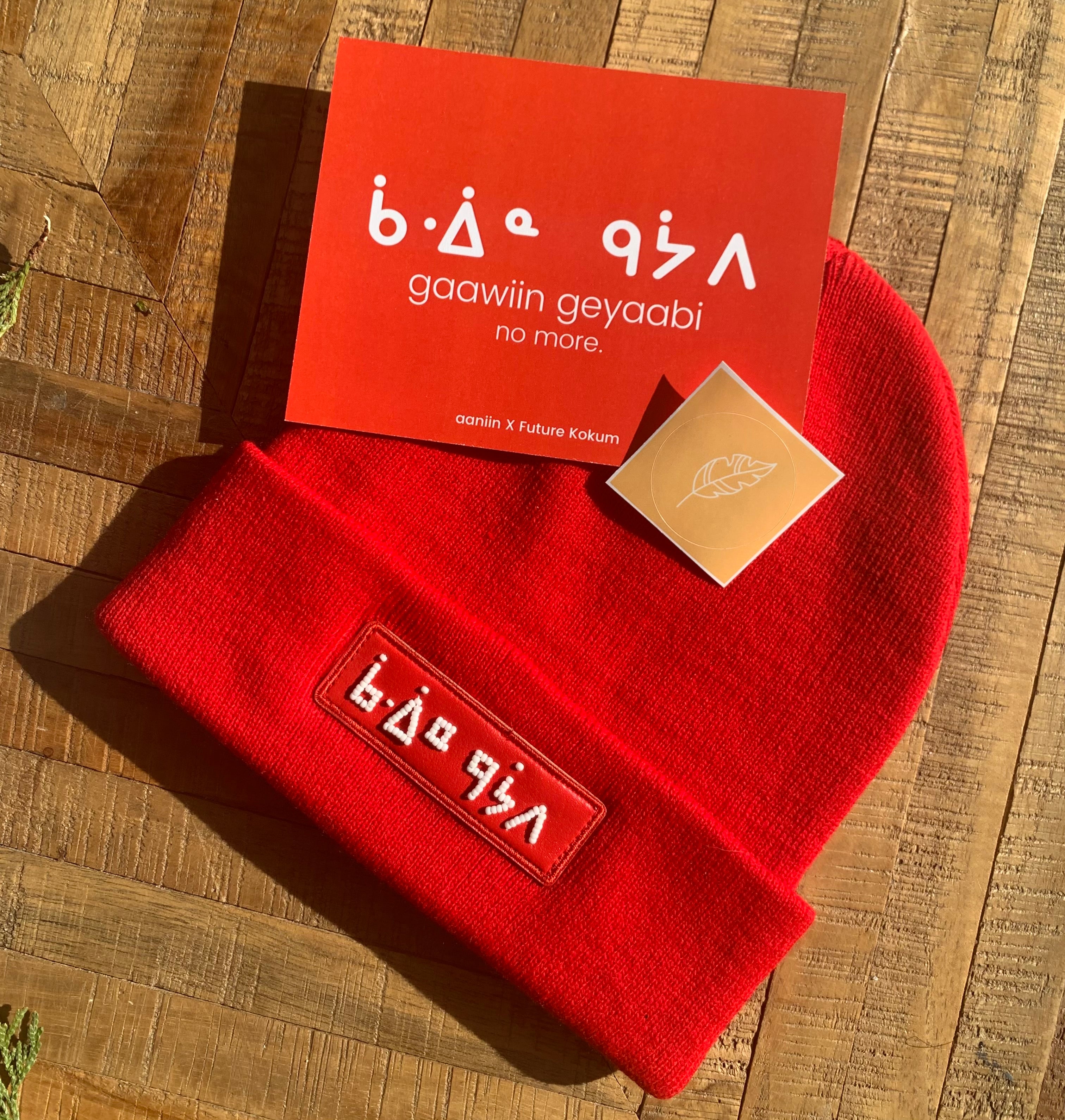A red beanie with beaded patches, placed on a wooden surface, with an information card beside it.