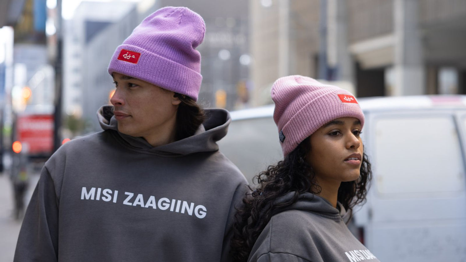 Two people wearing pink/purple aaniin beanies and gray 'more than land' hoodies with visible text in an urban setting of Toronto by Kyle Benjamin Turner.