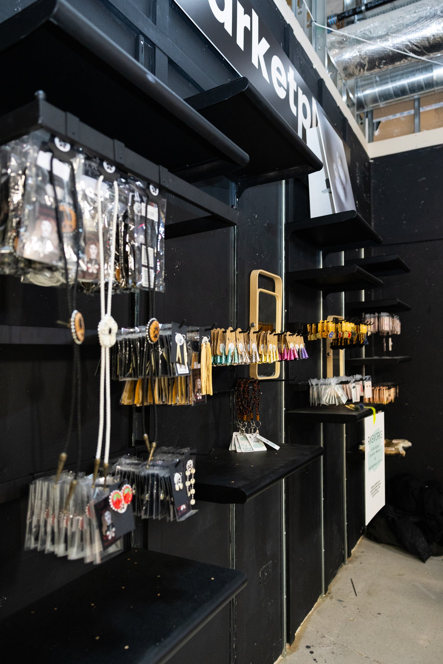 Black shelving unit with bimaadiziwin marketplace vendors on display in aaniin retail inc store. Vendors featured include Indigenous Bijoux, Riksworks, and our wholesale brand Dowan Winyan.