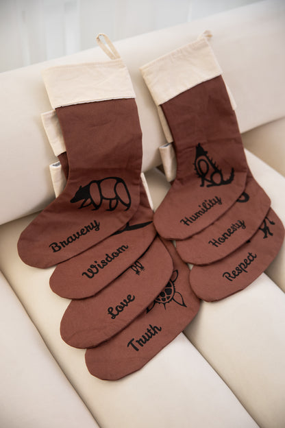 Brown Christmas stockings with embroidered text on a white surface. The Embroidered text displays a woodland cultural entity next to one of seven Indigenous grandfather teachings.