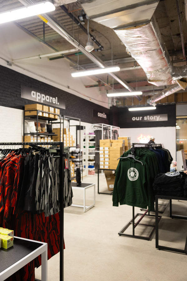 Aaniin Apparel store interior with clothing racks featuring the Aaniin Collection, Lesley Hampton collection, and Manitobah Moccassin and Mukluk's. In the foreground we see a black ceiling perimeter with an 'apparel', 'aaniin', and 'our story.' sign. Photo by Benjamin's Agency Inc.