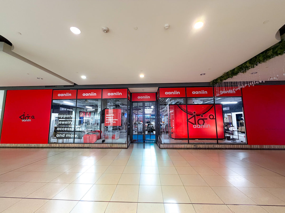 the Aaniin pop-up store location in the CF Toronto Eaton Centre between the Hoka store and Telus Mobile.  On the left we see the logo in black syllabics with white text. Beside it on the right we see the three north windows. The first window showing banners 'Apparel.' 'Jewelry.' 'Accessories.' 'and more.' with the third window displaying a red promotional poster. In the middle are metal french style doors. On the opposite side we see three south windows, and in the window a huge Aaniin shopping bag.
