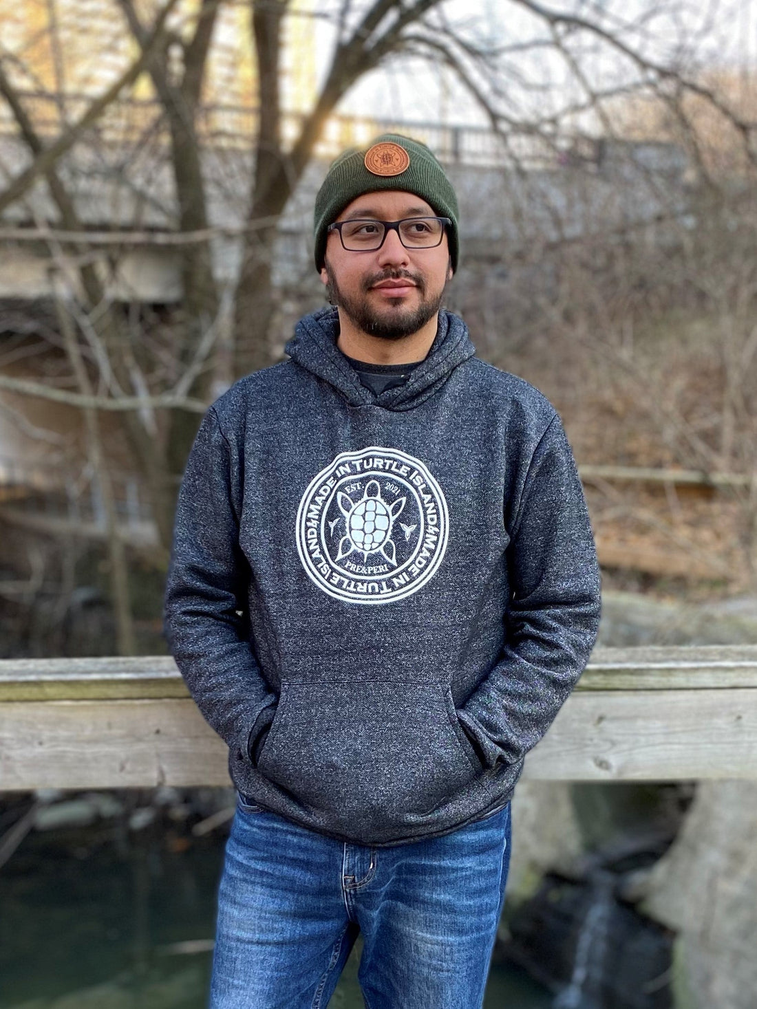 A bearded man wearing glasses, a green toque with tan leather patch, and salt and pepper hoodie with white screen printed graphic featuring a turtle with the words &quot;Made in Turtle Island&quot; around it