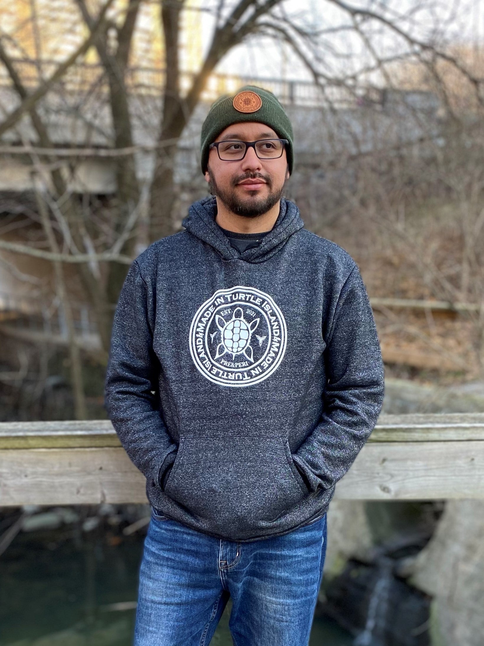 A bearded man wearing glasses, a green toque with tan leather patch, and salt and pepper hoodie with white screen printed graphic featuring a turtle with the words &quot;Made in Turtle Island&quot; around it