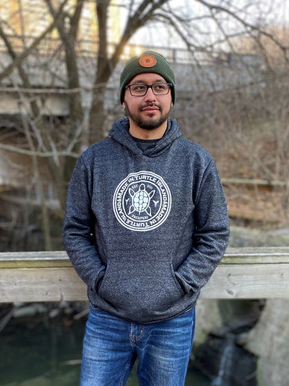 A bearded man wearing glasses, a green toque with tan leather patch, and salt and pepper hoodie with white screen printed graphic featuring a turtle with the words &quot;Made in Turtle Island&quot; around it
