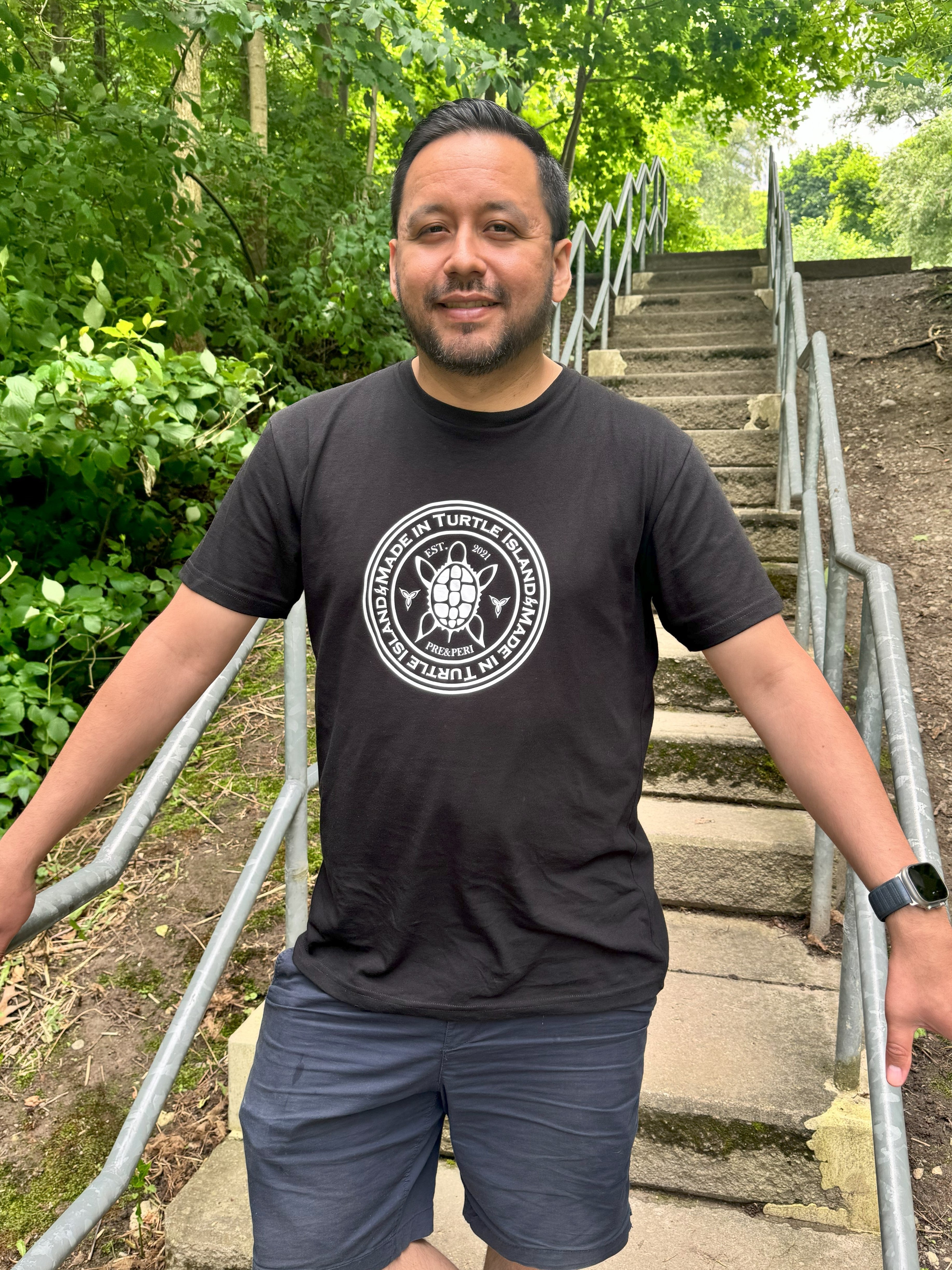 A man standing on outdoor steps wearing a black organic cotton t-shirt with a white print that says &quot;Made in Turtle Island&quot; around a central image of a turtle.