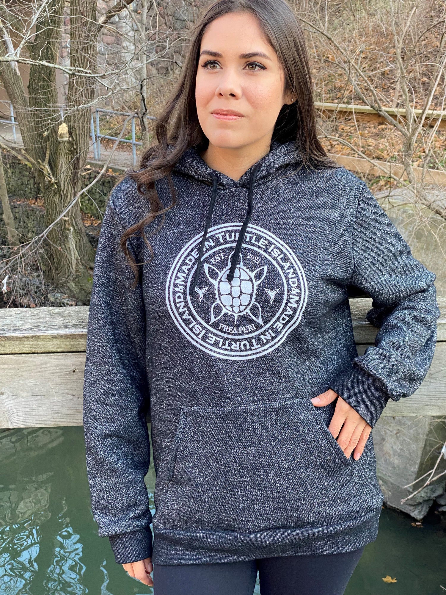 A woman wearing a salt and pepper hoodie with a white graphic featuring a stylized turtle with the words &quot;Made in Turtle Island&quot; around it.