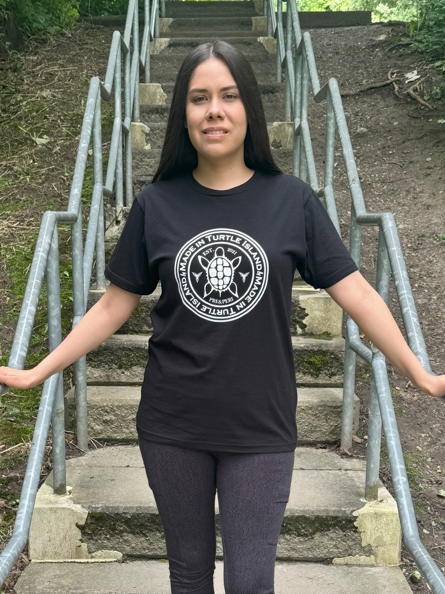 A woman standing on outdoor steps wearing a black organic cotton t-shirt with a white print that says &quot;Made in Turtle Island&quot; around a central image of a turtle.
