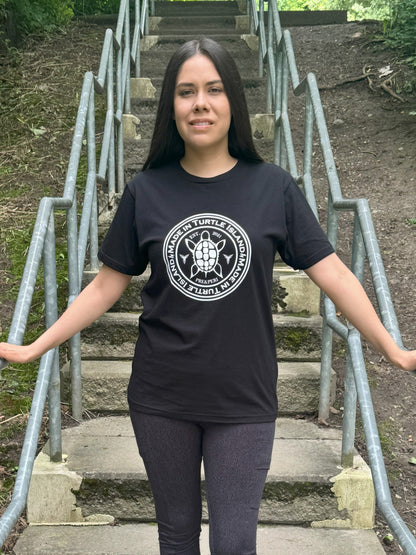 A woman standing on outdoor steps wearing a black organic cotton t-shirt with a white print that says &quot;Made in Turtle Island&quot; around a central image of a turtle.