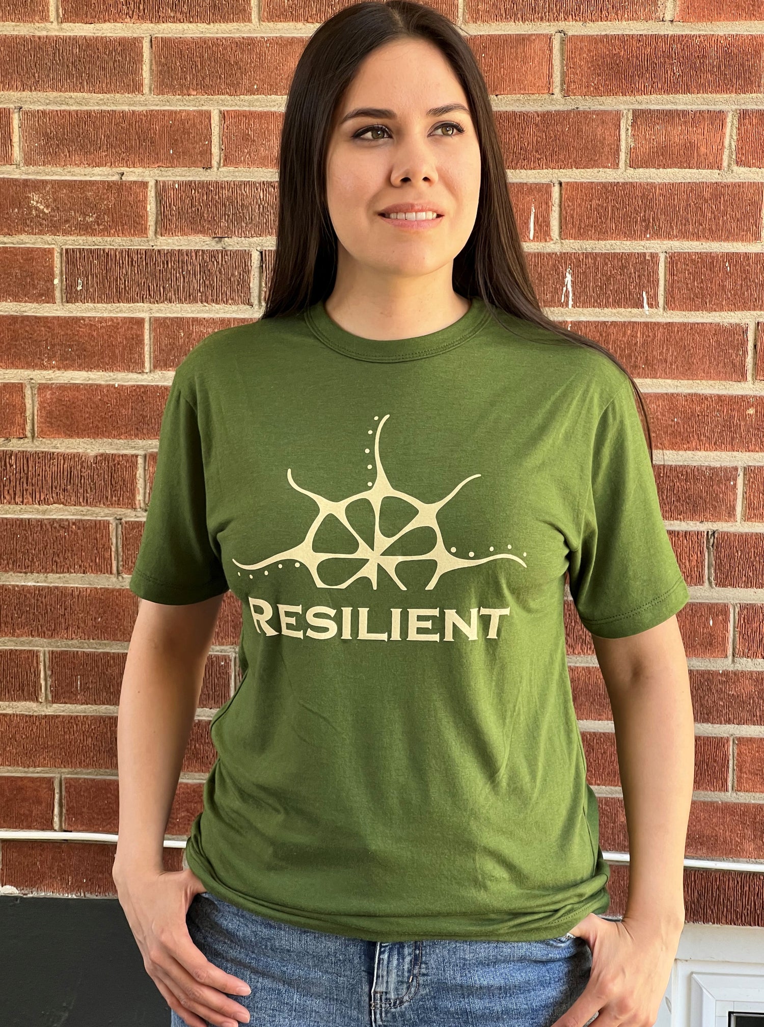 A woman wearing a green t-shirt with a pale yellow print that says &quot;Resilient&quot; under a stylized rising sun.