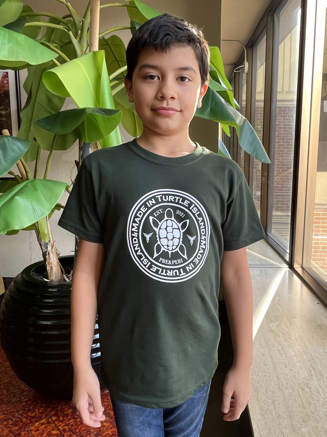 A boy wearing a forest green t-shirt with a white print of a turtle in a circle with the words &quot;Made in Turtle Island&quot; around it.