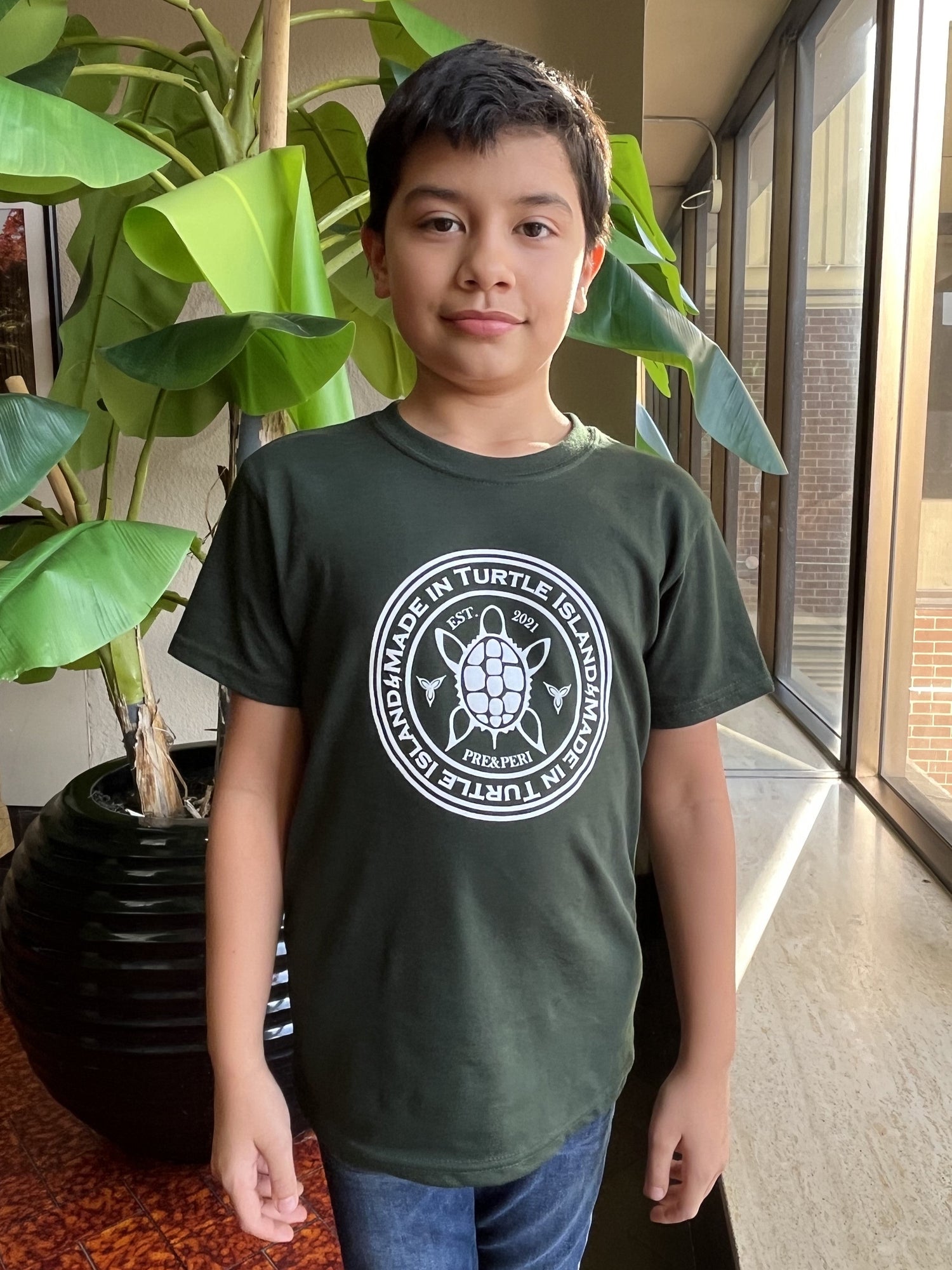 A boy wearing a forest green t-shirt with a white print of a turtle in a circle with the words &quot;Made in Turtle Island&quot; around it.
