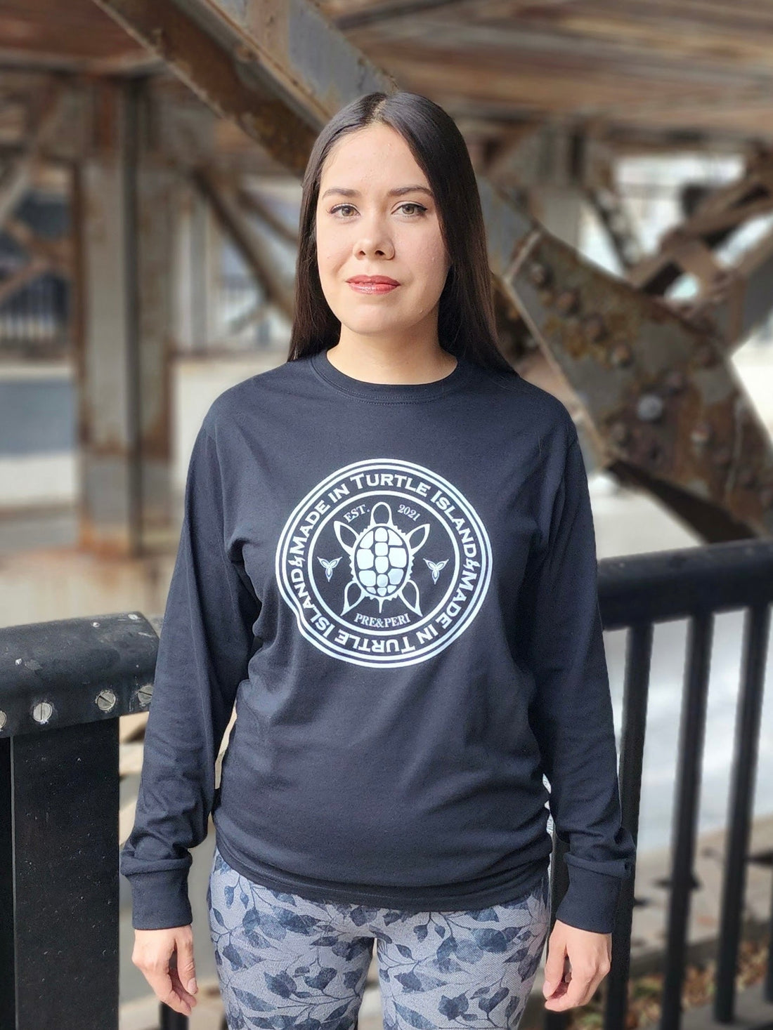 A woman wearing a black long sleeve t-shirt with a white print of a turtle in the centre of a circle with words &quot;Made in Turtle Island&quot; around it twice