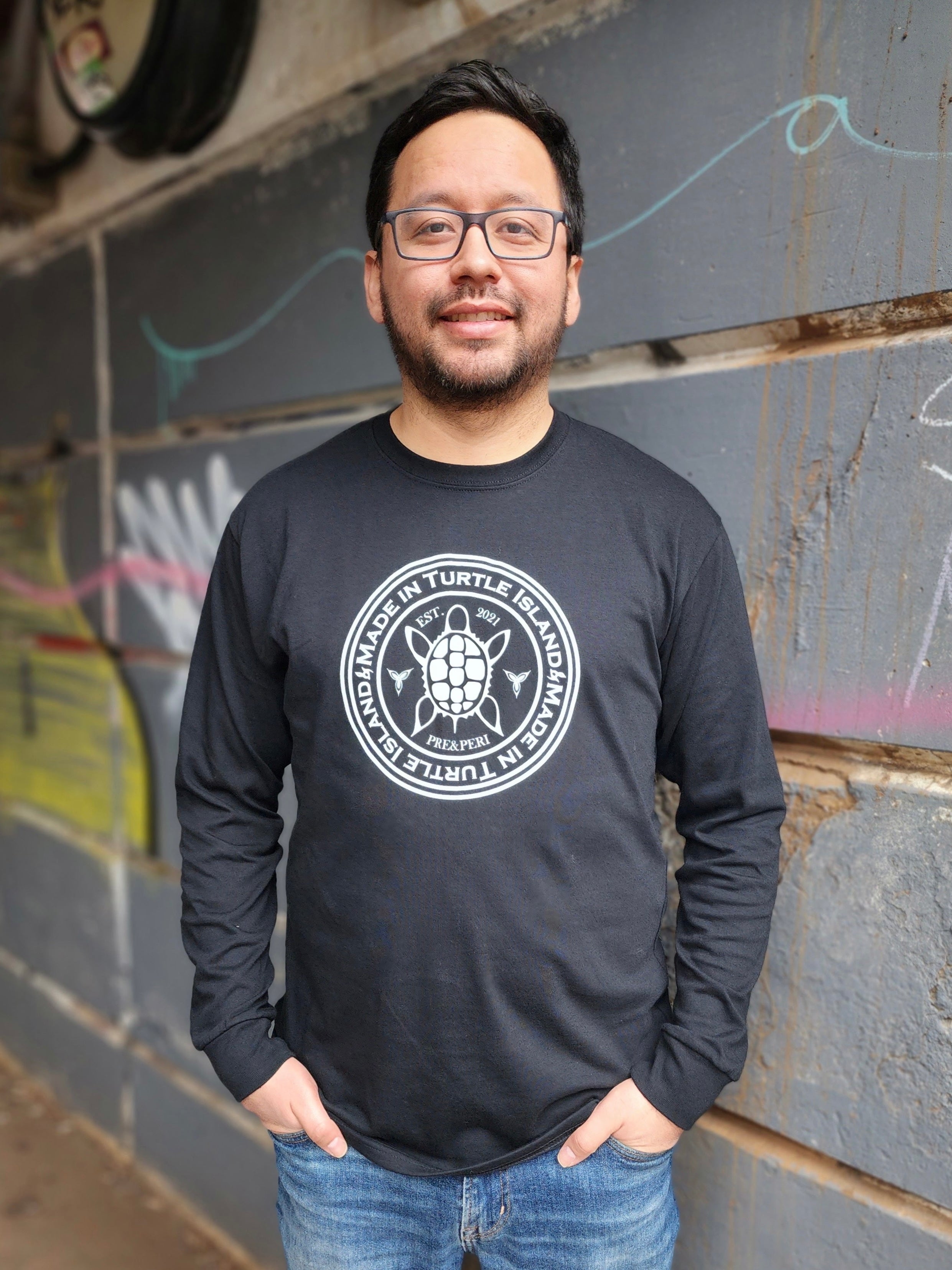 A man wearing a black long sleeve t-shirt with a white print of a turtle in the centre of a circle with words &quot;Made in Turtle Island&quot; around it twice