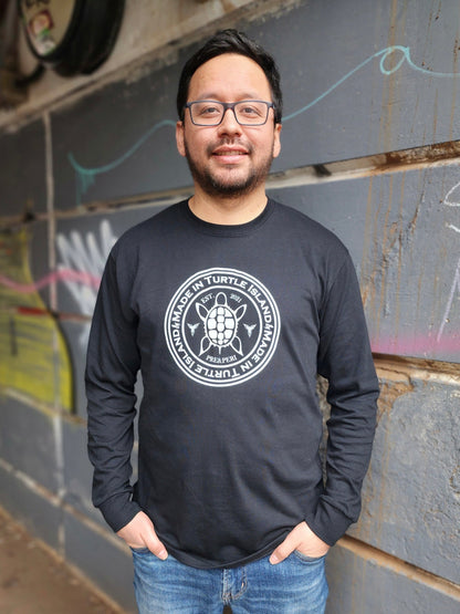 A man wearing a black long sleeve t-shirt with a white print of a turtle in the centre of a circle with words &quot;Made in Turtle Island&quot; around it twice